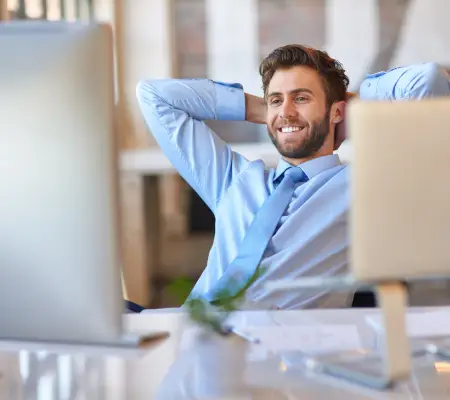 A smiling man in a blue dress shirt and tie leans back at his desk with his hands behind his head, relaxed about his home sale.